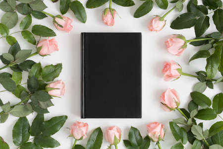Top view of a black book mockup surrounded by pink roses on a white table.の写真素材