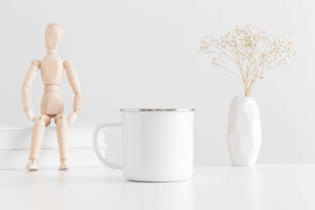 Enamel mug mockup with books, manikin and a gypsophila in a vase on a white table.の写真素材