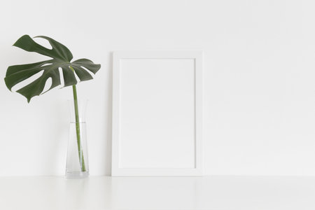 White frame mockup with a monstera leaf in a glass vase on a white table. Portrait orientation.