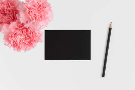 Top view of a black card mockup with a bouquet of pink carnations and workspace accessories on a white table.の写真素材