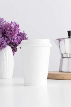 Coffee paper cup mockup with a moka pot and a bouquet of lilac in a vase on a white table.の写真素材