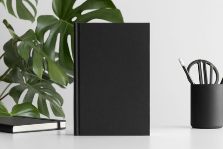 Black book mockup with workspace accessories on a white table and a monstera plant.の写真素材