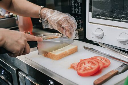 Woman hand cutting slice of homemade bread with slice fresh tomato on cutting knife in kitchen.Bakery products, freshly baked golden bread, cut pieces, millet,Prepare for making sandwich or salad.の写真素材