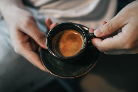 Man in white t-shirt hand holding a hot espresso in black ceramic cup and fresh homemade French croissant on wooden table. Breakfast with coffee and bread.Baked goods in traditional bakery.の写真素材
