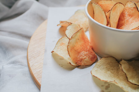 Fresh homemade deep fried crispy potato chips in white box on a wooden tray, top view. Salty crisps...