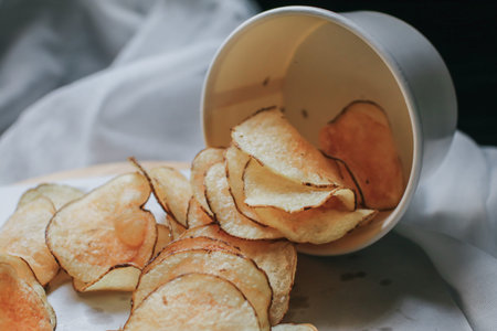 Fresh homemade deep fried crispy potato chips in white box on a wooden tray, top view. Salty crisps...