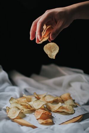Hand drop pieces of fresh homemade deep fried crispy potato chips on a wooden tray, top view. Salty crisps scattered on a table for a tasty snack break and party movie time.の写真素材