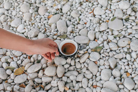 Hand pick up a black cup of hot coffee on white rock in garden floor. Background and texture of white pebbles stone. Take a break from work.の写真素材
