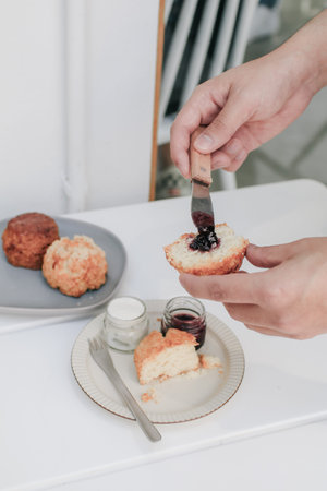 Hand spreads English Scones with Cream Cheese and Strawberry Jam, A scone is a single-serving quick bread, usually made of wheat, barley or oatmeal with baking powder.の写真素材