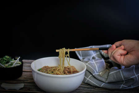 Hand uses chopsticks to pickup tasty egg noodle with pork and vegetable in five spices black soup on wooden table top view- Asian food style.の写真素材