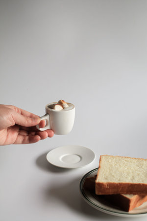 Hand holding cup of coffee with marshmallow and slice bread on white background. Morning breakfast, coffee break. Tasty beverage and sweet bakery. Winter and Autumn  conceptの写真素材