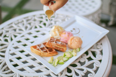 Hand pouring maple syrup on waffle and ice cream with apple on white table in garden. Tea time situation in bright mood background.  Healthy food conceptの写真素材