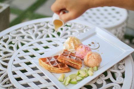 Hand pouring maple syrup on waffle and ice cream with apple on white table in garden. Tea time situation in bright mood background.  Healthy food conceptの写真素材