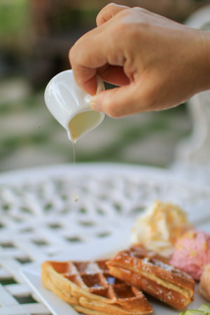 Hand pouring maple syrup on waffle and ice cream with apple on white table in garden. Tea time...