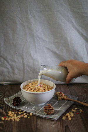 Hand pouring milk into granola bowl on wooden table with dark background. Healthy cereal and fiber...