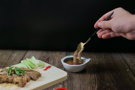 Hand with folk eating Grilled Pork Neck dipping spicy sauce on wooden board, Popular Appetizer in Thailand.の写真素材