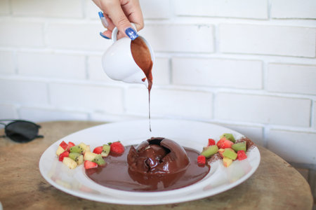 Pouring chocolate sauce over chocolate dome decorated with strawberry and kiwi in white plate....