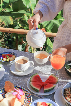 Woman hand pouring hot tea in white cup on wooden table set breakfast food outdoor in resort. Healthy food.の写真素材