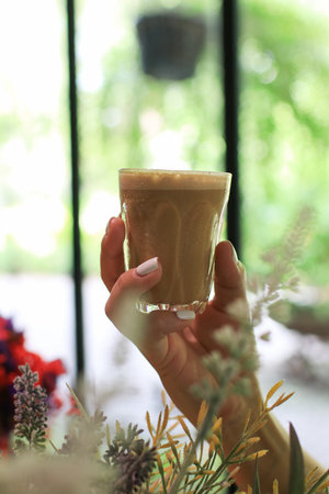 Hand holding glass of hot coffee latte with flower in vase on background. Morning Breakfast time. Vintage tone atmosphere.の写真素材