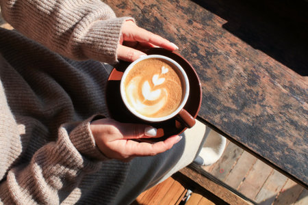 Hand holding hot coffee with latte art on wooden table under sunlight. Autumn Morning breakfast. Creamy coffee cup aromatic.の写真素材