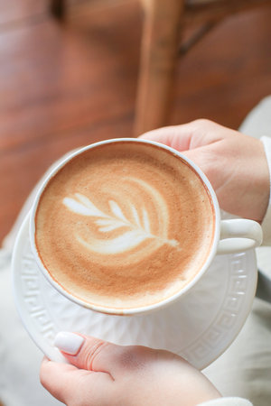 Hand holding hot coffee with foam milk latte art in white ceramic cup on wooden table. Autumn winter morning.の写真素材
