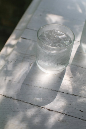Glass of iced water with aesthetic sunlight shadows on white wooden table. Healthy summer beverage, copy space.の写真素材