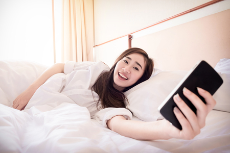Relaxed woman at home reading a text message on smartphone in her bright bedroom.の写真素材