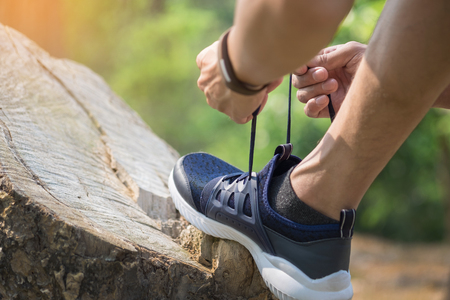 Cropped shot of young man runner tightening running shoe laces, getting ready for jogging exercise outdoors. Male jogger lacing his sneakers standing on forest path before morning run.の写真素材
