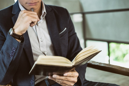 Thoughtful young business man in casual shirt holding note pad  in a notebook on a wooden table with pen working in a cafe.の写真素材