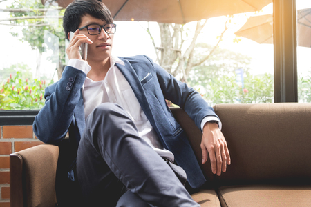 young asian businessman sitting relaxed on sofa making cellphone about his workの写真素材