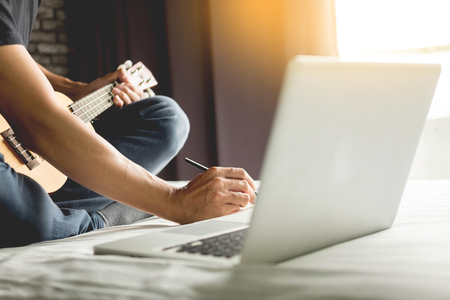 Happy young asian man playing ukulele sitting on bed in bedroomの写真素材