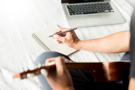 Happy young asian man playing ukulele sitting on bed in bedroomの写真素材