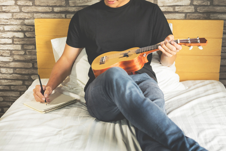 Happy young asian man playing ukulele sitting on bed in bedroomの写真素材
