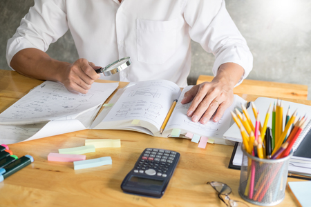 teenage man sitting by wooden table reading and writting on notebook in a classroom in fornt of a board with formulasの写真素材