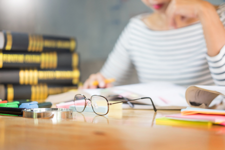 Charming girl sitting by wooden table reading and writting on notebook in a classroom in fornt of a board with formulasの写真素材