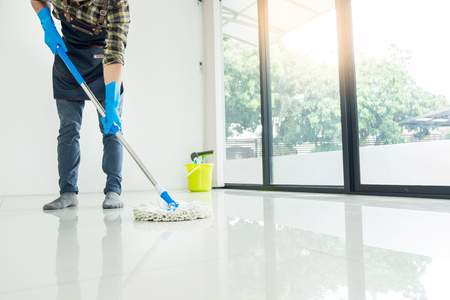 Young housekeeper cleaning floor mobbing holding mop and plastic bucket with brushes, gloves and detergents in the leaving room house floor helping his wifeの写真素材