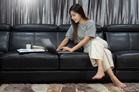 Pretty asian woman working remotely from home using laptop sitting on the couch or sofa in living room for work online with pet puppy cute dog and guardian, work life balance conceptの写真素材