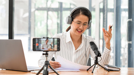 Asian women podcaster podcasting and recording online talk show at studio using headphones,...