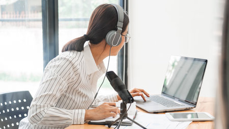 Asian women podcaster podcasting and recording online talk show at studio using headphones,...