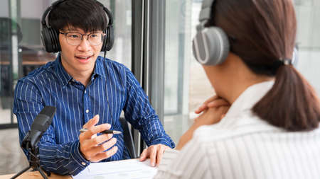 Asian woman radio hosts gesturing to microphone while interviewing a man guest in a studio while recording podcast for online show in studio together.の写真素材