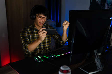 Excited asian man pro gamer sitting at the table, playing and Winning in online video games on a computer and smartphone, technology gaming cyber or e-sport championship concept.の写真素材
