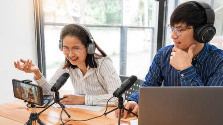 Asian Man and woman podcasters in headphones recording content with colleague talking to microphone and camera in broadcast studio together, communication technology and entertainment conceptの写真素材