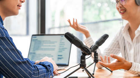 Asian woman radio hosts gesturing to microphone while interviewing a man guest in a studio while recording podcast for online show in studio together.の写真素材