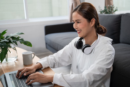 Happy casual beautiful Asian woman working on a laptop while sitting on sofa in living room at her...