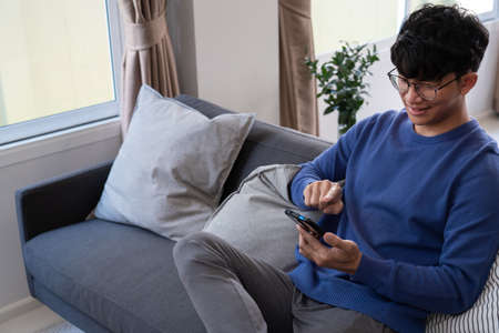 Handsome Asian teenager man wearing casual clothes in glasses using smart phone browsing Internet on mobile phone device at the living roomの写真素材
