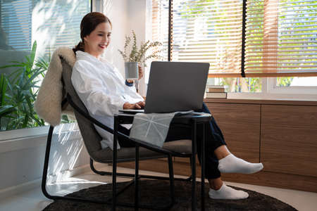 Happy casual beautiful Asian woman working on a laptop while sitting on chair in living room at her homeの写真素材