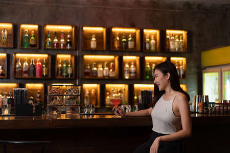 Nightlife concept a young woman in sexy outfits holding a glass of pink liquid enjoying its taste among the dim light in the bar.の写真素材