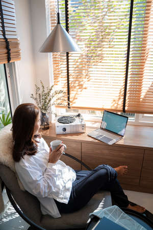 Happy casual beautiful Asian woman working on a laptop while sitting on chair in living room at her homeの写真素材