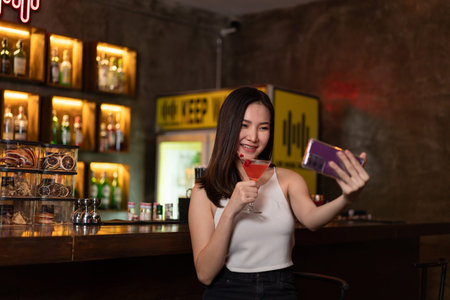 Nightlife concept a sexy girl with smiling face doing selfie on her smartphone while hold a vivid pink champaign in the night bar.の写真素材