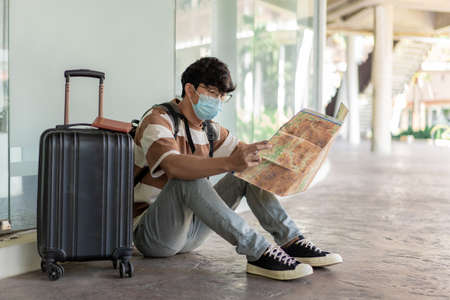 Travel, A man wearing glasses with a striped shirt and mask sit on the floor, reading map in his hand with suitcase placed on right side and backpack.の写真素材
